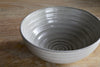 The nkuku Valinha Earthenware Cereal Bowl - Grey features a handmade, light gray spiral design with a smooth matte finish and unique color and texture variations, displayed on a wooden surface.