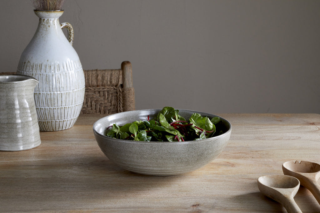The nkuku Valinha Earthenware Serving Bowl - Grey, featuring a textured spiral design, is filled with fresh greens on a wooden table beside wooden utensils and ceramic vessels, with a woven chair and neutral wall in the background.