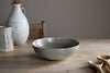 The Valinha Earthenware Serving Bowl by nkuku in grey features a reactive glaze and sits on a wooden table near wooden spoons, a ceramic pitcher, and a white vase, with a wicker chair in the background.
