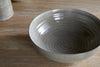 The Valinha Earthenware Serving Bowl by nkuku, featuring a large, round gray design with a subtle spiral texture, sits on a light wooden surface beside two other partially visible ceramic pieces.