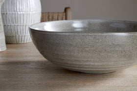 A close-up of the nkuku Valinha Earthenware Serving Bowl in grey, featuring a textured spiral design, sits on a wooden table with a white vase and woven chair softly visible in the background.