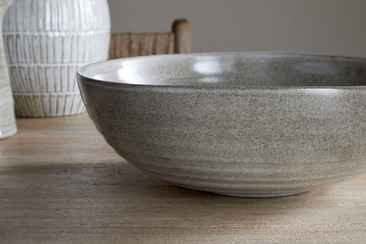 A close-up of the nkuku Valinha Earthenware Serving Bowl in grey, featuring a textured spiral design, sits on a wooden table with a white vase and woven chair softly visible in the background.