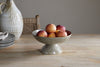 The nkuku Valinha Earthenware Footed Bowl in grey, filled with peaches, sits on a wooden table. A woven chair, stacked plates, and a tall textured ceramic pitcher complete the rustic tablescape.