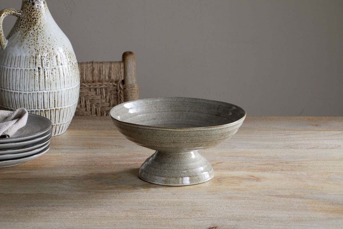 The nkuku Valinha Earthenware Footed Bowl in grey, with its speckled finish, rests on a wooden table beside plates, a napkin, and a jug—ideal for a rustic tablescape. A woven chair is visible in the background.
