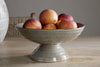 A Valinha Earthenware Footed Bowl - Grey by nkuku, filled with five ripe peaches, sits on a wooden table with a wicker chair and white vase in the background for a rustic tablescape.