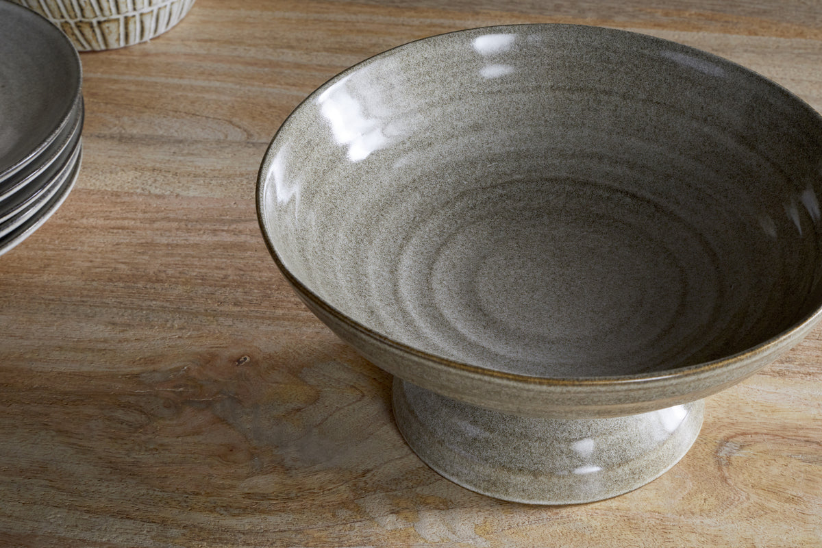 The nkuku Valinha Earthenware Footed Bowl - Grey, featuring a wide, shallow design and ribbed texture, adds rustic charm to any table setting. Shown on wood beside matching plates.