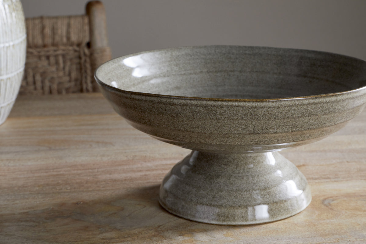 The Valinha Earthenware Footed Bowl - Grey by nkuku sits on a wooden table, with a woven chair and white vase in the background—ideal for creating a rustic tablescape.
