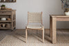 The nkuku Vinay Woven Armless Dining Chair in Natural, featuring a jute seat and back, stands on a patterned rug between an acacia wood dining table and sideboard in a rustic, neutral room.