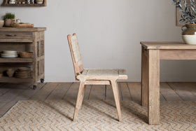 The Vinay Woven Armless Dining Chair by nkuku, made of acacia wood with a woven jute seat and backrest, is placed next to a wooden dining table in a rustic kitchen with wooden cabinets, shelves, and neutral decor.