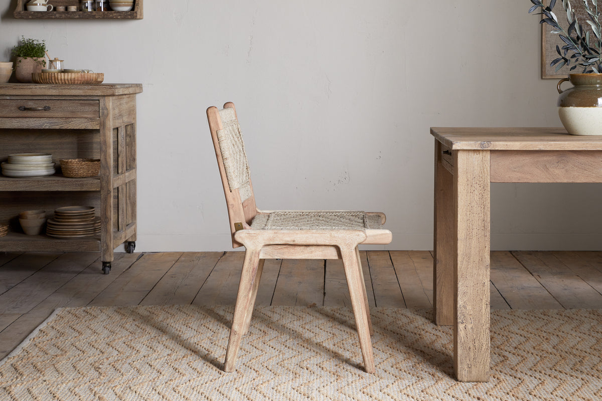 The Vinay Woven Armless Dining Chair by nkuku, made of acacia wood with a woven jute seat and backrest, is placed next to a wooden dining table in a rustic kitchen with wooden cabinets, shelves, and neutral decor.
