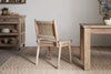 The nkuku Vinay Woven Armless Dining Chair in natural jute sits on a patterned rug in a rustic dining room with acacia wood furniture and neutral decor.