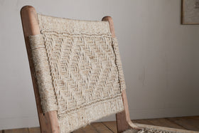 The Vinay Woven Armless Dining Chair by nkuku features acacia wood and a geometric diamond-patterned jute backrest. Set against a light wall and wooden floor, its natural style stands out in any dining space.