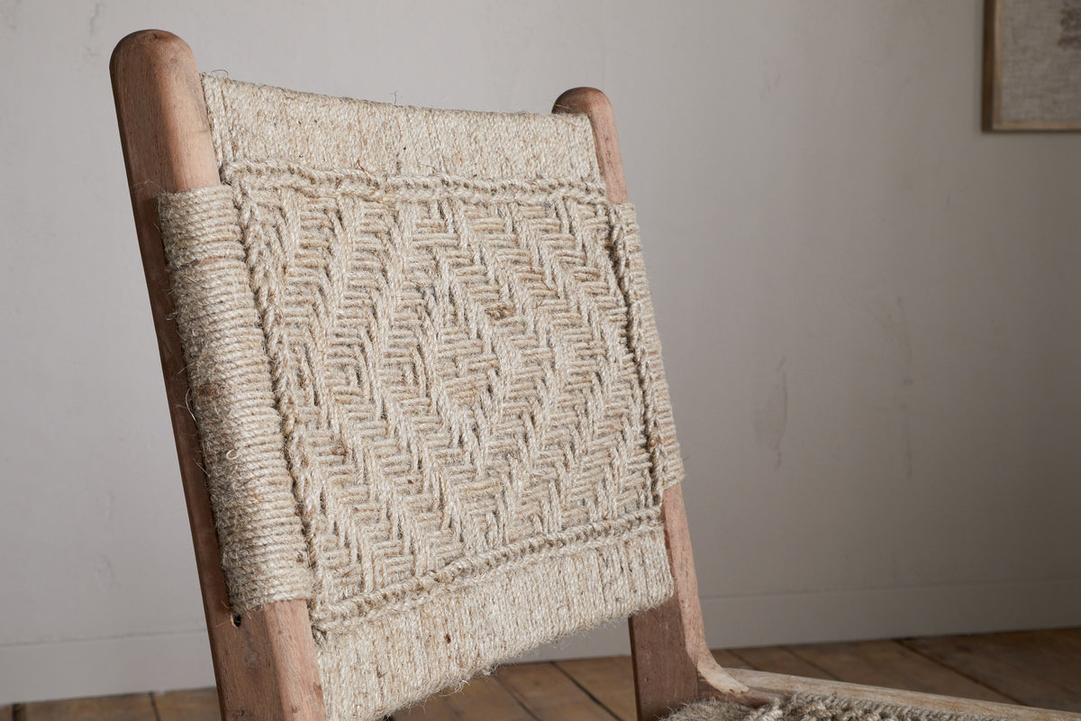 The Vinay Woven Armless Dining Chair by nkuku features acacia wood and a geometric diamond-patterned jute backrest. Set against a light wall and wooden floor, its natural style stands out in any dining space.