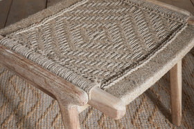 A close-up of the Vinay Woven Armless Dining Chair by nkuku, crafted from acacia wood with a geometric diamond-patterned jute seat, placed on a textured rug with a matching design.
