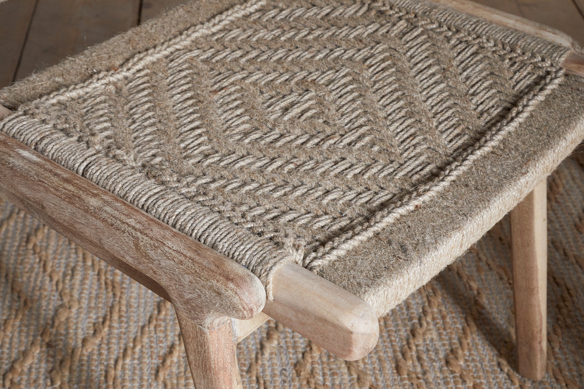 A close-up of the Vinay Woven Armless Dining Chair by nkuku, crafted from acacia wood with a geometric diamond-patterned jute seat, placed on a textured rug with a matching design.