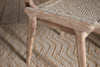 The Vinay Woven Armless Dining Chair by nkuku, featuring a woven seat and acacia wood frame in a natural finish, sits atop a patterned woven jute rug on a wooden floor, creating an inviting rustic look.