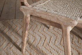 The Vinay Woven Armless Dining Chair by nkuku, featuring a woven seat and acacia wood frame in a natural finish, sits atop a patterned woven jute rug on a wooden floor, creating an inviting rustic look.
