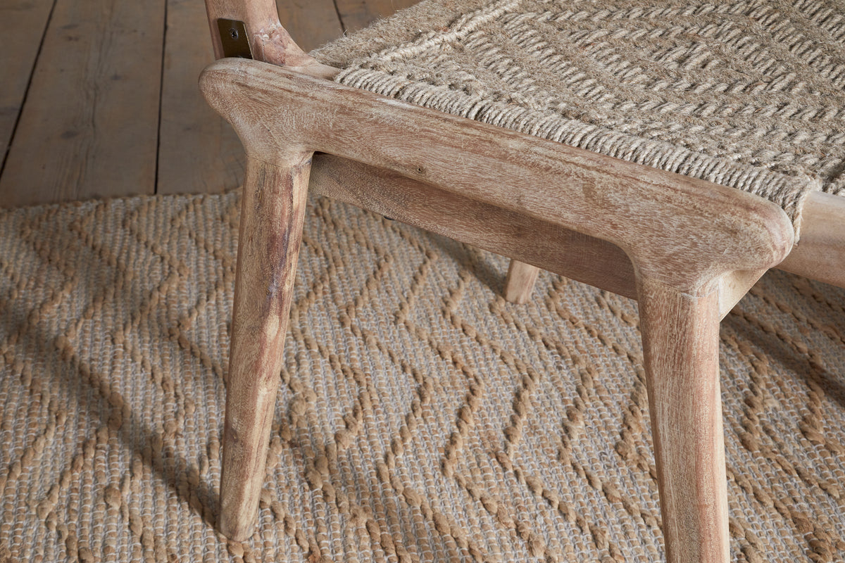 The Vinay Woven Armless Dining Chair by nkuku, featuring a woven seat and acacia wood frame in a natural finish, sits atop a patterned woven jute rug on a wooden floor, creating an inviting rustic look.