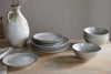 The nkuku Valinha Earthenware Dinnerware Set - Grey (Set of 12) features ceramic plates and bowls with a gray speckled glaze, arranged on a wooden table beside a cream pitcher and a folded napkin.