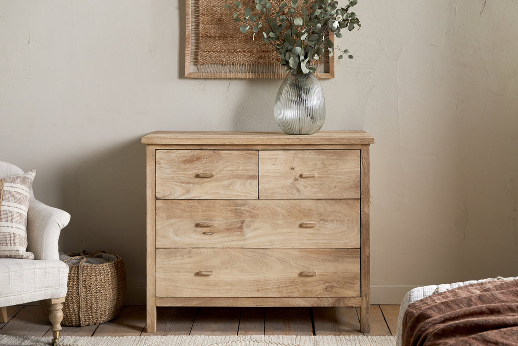 The Vivan Grooved Chest of Drawers - Natural by nkuku stands against a beige wall, topped with a clear textured vase holding greenery. An armchair with a striped cushion and woven basket sits to the left; part of a bed is visible on the right.