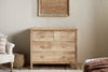 The nkuku Vivan Grooved Chest of Drawers in natural wood stands against a beige wall beneath a woven wall hanging, with part of a white chair, a woven basket, and a bed with a brown blanket visible nearby.