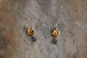 The nkuku Veera Labradorite Earrings - Gold feature textured, round hammered metal with dark grey stone drops, displayed on a rough, grayish-brown stone surface.