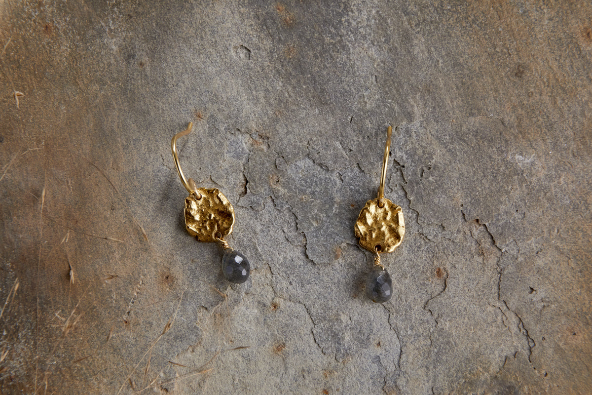 The nkuku Veera Labradorite Earrings - Gold feature textured, round hammered metal with dark grey stone drops, displayed on a rough, grayish-brown stone surface.