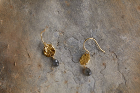 The nkuku Veera Labradorite Earrings - Gold feature hammered, irregular oval shapes and dark labradorite stone drops, shown on a rough gray stone surface.