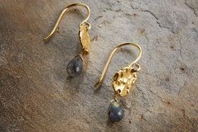 The Veera Labradorite Earrings – Gold by nkuku feature gold-coloured hooks with textured oval details and faceted labradorite drops, shown on a stone surface.