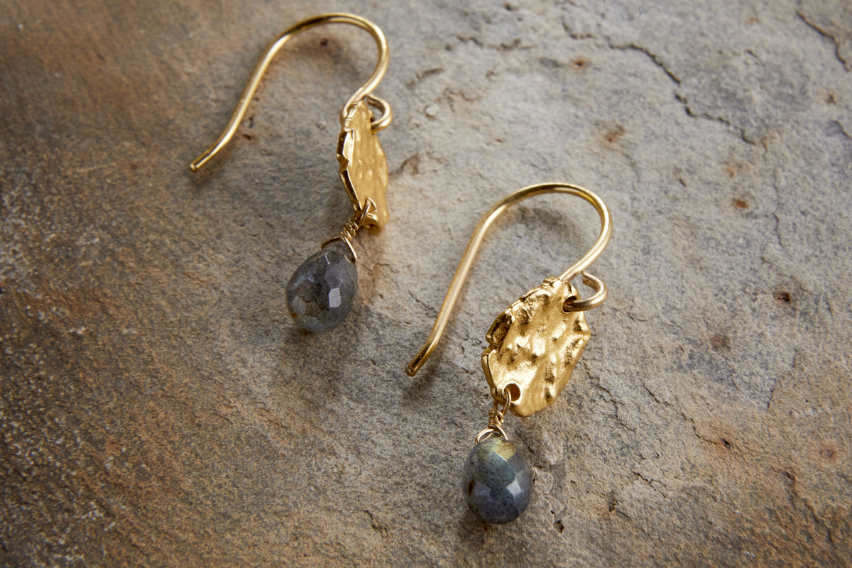 The Veera Labradorite Earrings – Gold by nkuku feature gold-coloured hooks with textured oval details and faceted labradorite drops, shown on a stone surface.