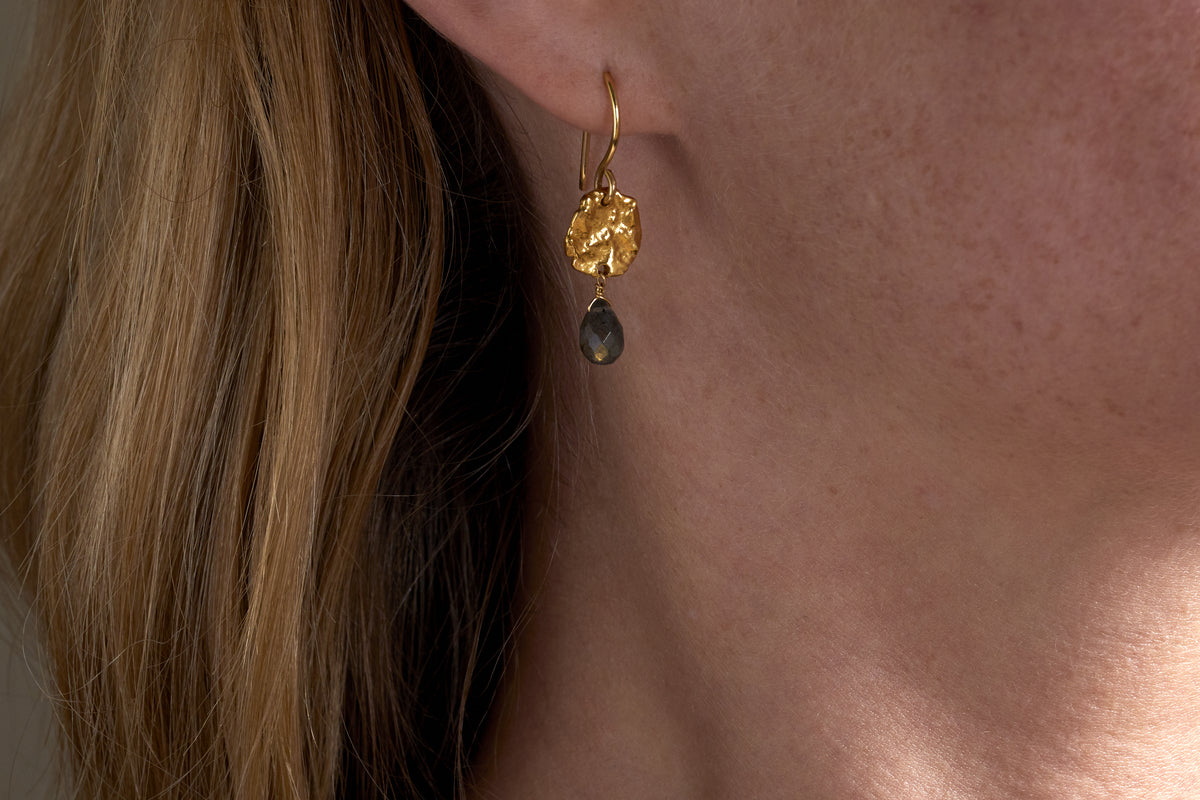 Close-up of a womans ear wearing nkukus Veera Labradorite Earrings - Gold, featuring a textured round gold top and a dark teardrop-shaped labradorite stone. Light brown hair is partially visible.