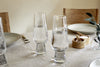 Two clear Visla Champagne Flutes by nkuku are filled and placed on a beige tablecloth. The table features light gray plates, wooden chairs, a fork, a small wooden salt cellar, and a vase with green leaves.