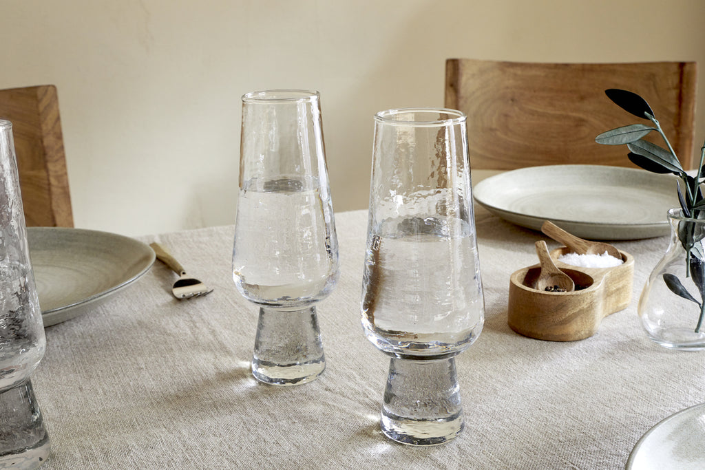 Two clear Visla Champagne Flutes by nkuku are filled and placed on a beige tablecloth. The table features light gray plates, wooden chairs, a fork, a small wooden salt cellar, and a vase with green leaves.