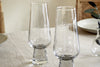 Four clear Visla Champagne Flutes by nkuku sit on a light tablecloth, accompanied by plates and a small vase with leaves in the background.