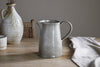 The Valinha Earthenware Jug in grey by nkuku rests on a wooden table beside a folded beige cloth, with a textured vase and ceramic cup in the background, creating a calm, rustic ambiance perfect for unique dining.