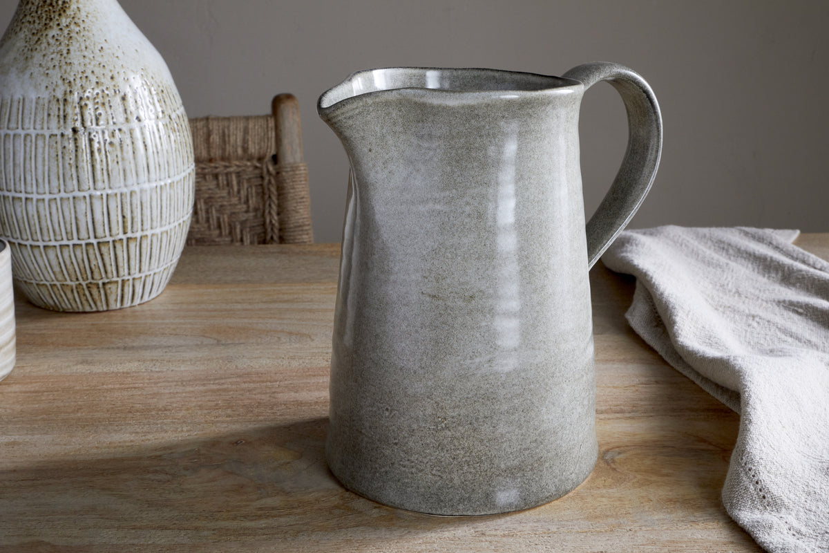 The Valinha Earthenware Jug in Grey (Large) from nkuku sits on a wooden table, bringing elegance to your serveware. Behind it, a textured vase and woven chair enhance the earthy decor, while a draped cloth softens the dining scene.