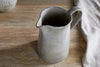 The Valinha Earthenware Jug - Grey - Large by nkuku rests on a wooden surface beside a textured white cloth. Its reactive glaze and elegant handle catch the soft natural light, making it ideal as elevated serveware for any dining experience.