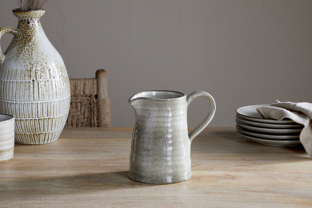 The Valinha Earthenware Jug - Grey - Small by nkuku rests on a wooden table, accompanied by stacked plates, a cloth, and a large textured vase. A woven chair completes this calm, rustic setting—ideal for an elevated dining experience.
