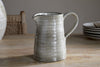 The Valinha Earthenware Jug - Grey - Small by nkuku, featuring a smooth, light gray reactive glaze, sits on a wooden table. A woven chair and stacked plates appear in the background, creating an elevated dining setting.