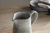 The nkuku Valinha Earthenware Jug in grey (small) and distinctive tableware adorn the table, enhancing your dining experience.