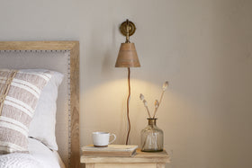 A cozy bedside features a soft beige bed, striped pillows, and a wooden table with a white cup on stacked books. The nkuku Vattu Mango Wood Plug In Wall Lamp in Natural & Antique Brass casts warm light over neutral walls beside a dried-stem vase.