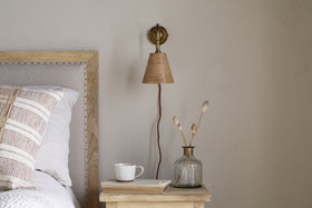 A cozy bedroom corner features a wooden bed, beige linens, and a nightstand with books, a cup, and dried flowers. Above, the nkuku Vattu Mango Wood Plug In Wall Lamp in Natural & Antique Brass provides country-style lighting on the light wall.