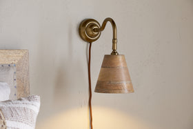 The nkuku Vattu Mango Wood Plug In Wall Lamp in Natural & Antique Brass is mounted on a beige wall, its brown cord hanging down as warm, ambient light glows. A bed with a textured pillow appears at the left edge.