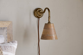 The nkuku Vattu Mango Wood Plug In Wall Lamp in Natural & Antique Brass is mounted on a beige wall, adding natural texture. A brown cord hangs beneath, and part of a wooden bed frame with a striped pillow appears at the edge.