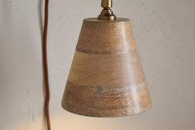 A close-up of the nkuku Vattu Mango Wood Plug In Wall Lamp in natural and antique brass shows its conical wooden shade, brown cord, and textured off-white wall backdrop. The lamp is unlit.