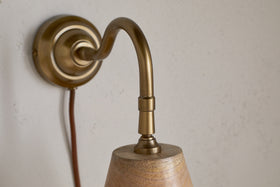 A close-up of the nkuku Vattu Mango Wood Plug In Wall Lamp in Natural & Antique Brass, featuring a conical wooden shade, brass curved arm, and brown fabric cord, mounted on a subtly textured beige wall.