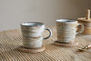 The nkuku Vizo Mugs - Grey (Set of 2) feature a unique two-tone reactive glaze and rest on wooden coasters atop a striped beige tablecloth, with a wooden container and spoon in the background highlighting their distinctive finish.