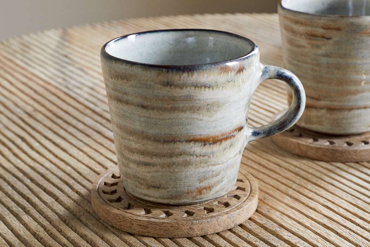 The nkuku Vizo Mugs - Grey (Set of 2) feature a distinctive marbled finish and rest on a decorative wooden coaster atop a ribbed table, with another stylish mug partially visible in the background.