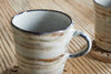 A Vizo Mug - Grey by nkuku, featuring a two-tone reactive glaze with marbled beige and brown, rests on a wooden coaster atop a light wood table. A second matching mug from the set is partially visible in the background.