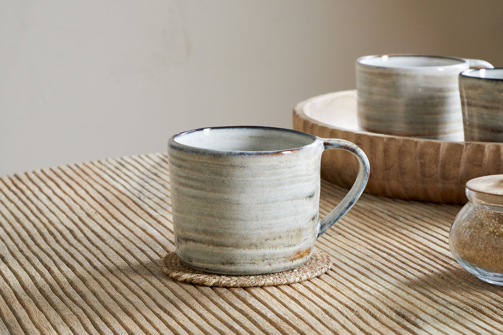 The nkuku Vizo Mug - Grey - Large sits on a round woven coaster atop a ribbed wooden table, with a tray of unique tableware and a small glass jar in the background.